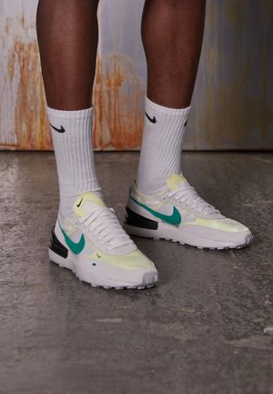 Osoba nosząca białe buty sportowe Nike z zielonym logo oraz białe skarpetki z logo Nike stoi na betonowej podłodze.