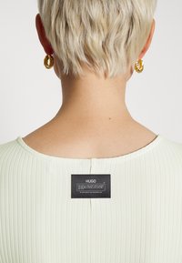 Mintgrön ribbad tygtröja med vid halsringning, med en svart varumärkesetikett på baksidan, och guldfärgade hoop örhängen synliga.