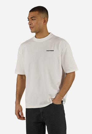 Mann trägt ein schlichtes weißes übergroßes T-Shirt mit dem Schriftzug "PEGADOR" auf der Brust und schwarze Hosen, steht mit einer Hand in der Tasche.