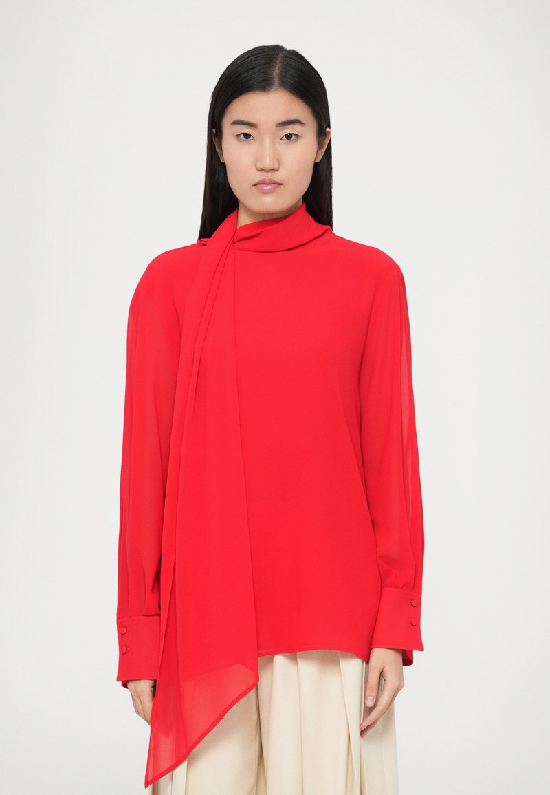 Blouse en chiffon rouge avec un col montant et une longue écharpe fluide. Manches longues avec détails à boutons. Texture lisse et légère.