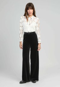 Femme debout, portant un blouse en dentelle blanche transparente et un pantalon noir à taille haute et coupe large, avec des chaussures noires sur un fond clair et uni.