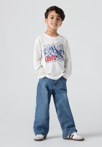 Bijela košulja s dugim rukavima s plavim grafičkim motivom i crvenim natpisom "Levi's", uparen s ležernim plavim trapericama; ležerni tenisice upotpunjuju izgled.