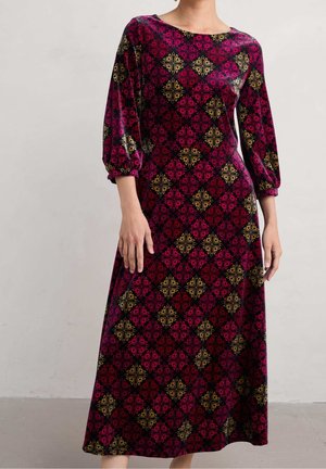 Femme portant une robe longue à manches longues avec un motif floral géométrique bordeaux et or sur fond noir.