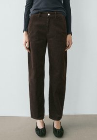 Pantalon en velours côtelé marron à coupe droite, avec fermeture à boutons sur le devant et poches, associé à des ballerines noires arrondies.