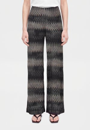 Une femme porte un haut blanc, une chemise bleu clair ouverte, un pantalon à jambes larges et à motifs ainsi que des sandales noires à talons, sur un fond uni.