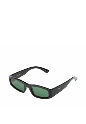 Lunettes de soleil rectangulaires noires avec des verres vert foncé et des montures épaisses, inclinées pour montrer la branche droite portant la marque Komono.