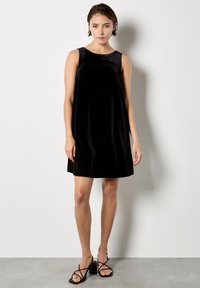 Czarna sukienka bez rękawów z miękkiego weluru, do kolan, z okrągłym dekoltem. Połączona z czarnymi sandałami na słupku z paskami.