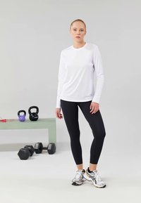 Valge pika varrukaga sportlik pluus, mis on kombineeritud mustade retuusidega. Mudel kannab jooksutossusid. Taustal on harjutustooted ja kettlebell'id heledal pinnal.