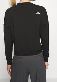 Persona che indossa una maglia a maniche lunghe nera di North Face e pantaloni grigio scuro, vista di spalle su uno sfondo bianco.