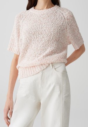 Femme portant un pull en maille rose clair à texture douce et manches courtes, rentré dans un pantalon taille haute blanc cassé.