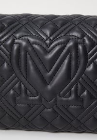 Černá prošívaná kožená taška s výrazným logem ve tvaru srdce. Texturovaný povrch s diamantovým vzorem a kontrastním prošitím.