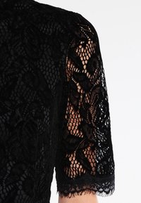 Svart spetsärm med ett blommönster, semi-genomskinlig textur och vågig nederkant, med ett fast svart tyg som bas under.