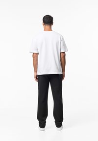 Bílá tričko s krátkým rukávem zobrazené zezadu, doplněné černými tepláky a bílými teniskami, na jednoduchém bílém pozadí.