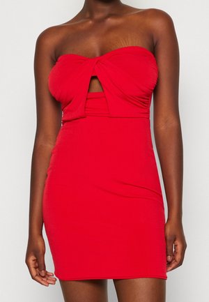 Mujer que lleva un mini vestido ajustado, sin tirantes, de color rojo brillante, con un detalle de nudo retorcido en el busto y un recorte debajo.