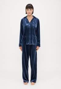 Conjunto de pijama em veludo de um azul marinho profundo. Parte de cima com botões, duas bolsos frontais, mangas longas e capuz. Calças largas com perna reta.