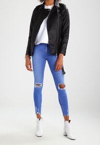 Chaqueta de cuero sintético negra con cremallera asimétrica, camiseta blanca, jeans ajustados desgastados azules y botas tobilleras blancas con acentos negros.