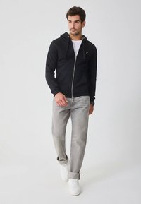 Felpa nera con zip e coulisse, con un piccolo logo sul petto, abbinata a jeans grigio chiaro e sneakers bianche.