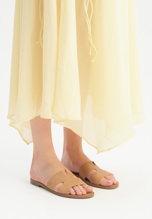 Bruine leren platte sandalen met uitsnijdingen, voorzien van een lage hak en een gestructureerde afwerking, gecombineerd met een lange, doorschijnende gele jurk.