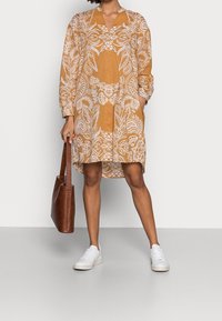 Langærmet, knælang kjole i sennepsgul med hvidt paisley-mønster, med V-hals; kombineret med hvide sneakers og en brun tote bag.