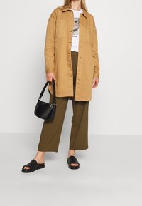 Ljusbrun skjortjacka med knappar, bärs över en t-shirt med tryck, matchad med olivgröna vida byxor och svarta slidesandaler. Svart crossbody-väska.