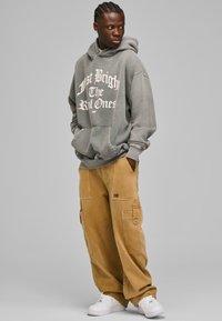 Grå hættetrøje med hvid tekst, løs pasform og frontlomme. Beige cargo bukser med flere lommer og broderet logo. Hvide sneakers.