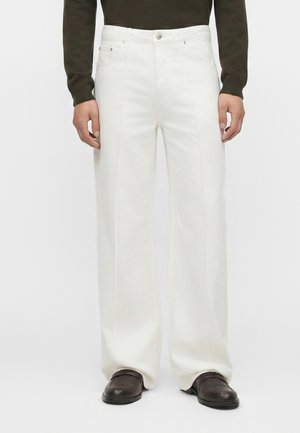 Άντρας που φοράει λευκό, ψηλόμεσο παντελόνι με φαρδιά μπατζάκια και μπροστινές ραφές, σε συνδυασμό με σκούρα μπλούζα και μαύρα παπούτσια, στέκεται μπροστά σε λευκό φόντο.