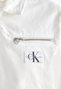 Tessuto in denim bianco con una tasca con zip argentata. Presenta un'etichetta Calvin Klein Jeans con il logo nero "CK".