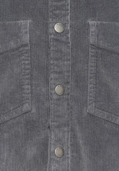 Grau gerippter Cordstoff mit vertikalen Streifen, zwei vorderen Taschen und drei metallenen Druckknöpfen, die vertikal in der Mitte angeordnet sind.