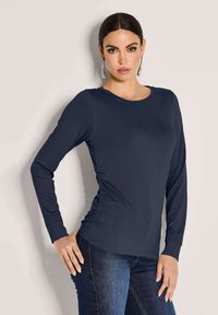 Vrouw met donker haar draagt een marineblauw longsleeve shirt en blauwe spijkerbroeken, staand tegen een effen lichte achtergrond met één hand op haar heup.