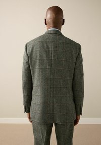 Man facing away wearing a gray plaid suit with brown lines, standing against a beige wall and carpeted floor.