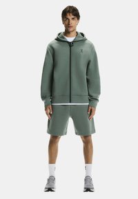 Groene fleece hoodie met ritssluiting en trekkoord, gecombineerd met bijpassende shorts. Heeft een ritssluiting aan de voorkant en een logo op de borst. Ademend materiaal.