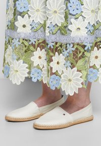 Chaussures espadrilles blanches portées avec une jupe en dentelle florale présentant de grandes fleurs brodées blanches et bleues avec des feuilles vertes.