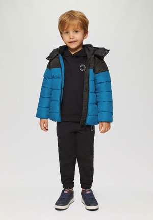 Blauwe en zwarte gewatteerde jas met een capuchon, gedragen over een zwarte hoodie en zwarte broek. Voorzien van een rits aan de voorkant en zijzakken. Casual sneakers.