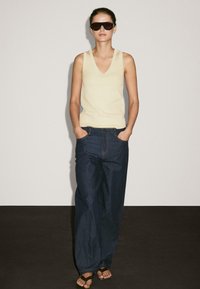 Lys gul ærmeløs top, mørkeblå vide jeans og mørke solbriller; står med hænderne i lommerne på en neutral baggrund.