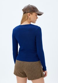 Donna che indossa un top a maniche lunghe azzurro aderente con coste, pantaloni corti marrone a spina di pesce, orecchini a cerchio dorati e un cappellino da baseball in pelle marrone, vista di profilo.