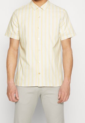 Mężczyzna ubrany w krótki rękaw, koszulę na guziki w pionowe żółte i białe paski, połączoną z jasnobeżowymi spodniami.