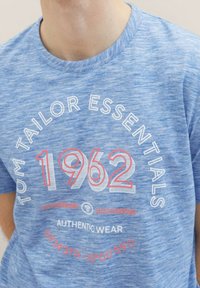 Modra melirana majica iz mehkega blaga. Spredaj je napis "TOM TAILOR ESSENTIALS" in "1962" v belih in koralnih črkah. Oblika z okroglim ovratnikom.