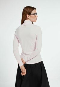 Jasnoróżowa bluzka z długim rękawem o dopasowanym kroju, z wysokim kołnierzem i małym złotym akcentem z tyłu, połączona z czarną spódnicą.