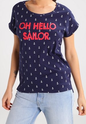 Granatowy t-shirt z białymi wzorami kotwic i wyraźnym czerwonym napisem "OH HELLO SAILOR", noszony z jasnoniebieskimi dżinsami.