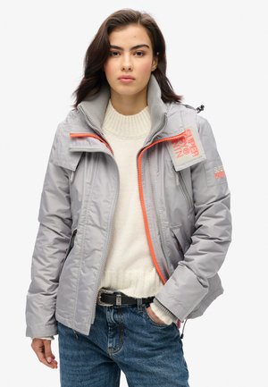 Veste matelassée gris clair avec un col montant, fermeture éclair orange contrastante et écussons logo sur la manche et la poitrine. Portée par-dessus un pull crème.