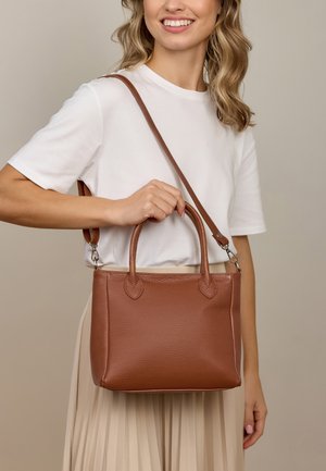 Frau in weißem Shirt und beiger Rock hält eine braune Lederhandtasche mit zwei Griffen und einem Schultergurt.