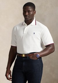 Bărbat purtând un tricou polo alb cu margini bleumarin și roșii, blugi închiși la culoare, curea maro și ceas de mână cu cadran albastru, stând cu o mână în buzunar.