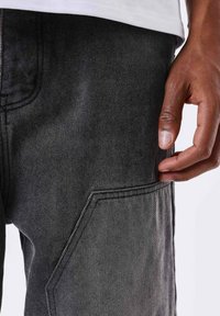 Pantaloni in denim nero con una finitura testurizzata, dotati di tasche laterali e cuciture visibili. Il tessuto è resistente e leggermente sbiadito.