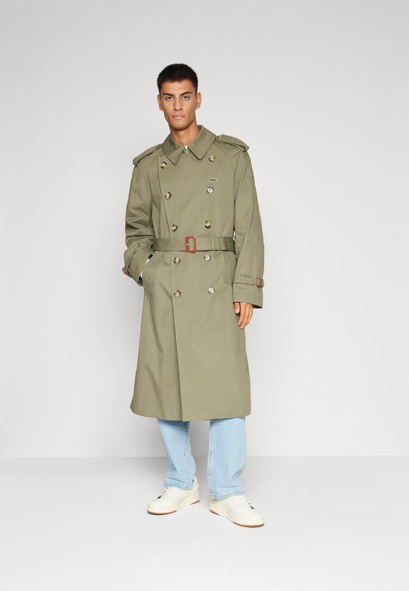 Mann trägt einen olivgrünen Gürtel-Trenchcoat, helle blaue Jeans und weiße Turnschuhe und steht vor einem schlichten hellen Hintergrund.