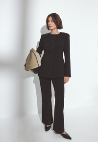 Svart dubbelknäppt blazer och högmidjade byxor med utsvängda ben, i kombination med en beige väska och spetsiga svarta skor, mot en vit vägg.