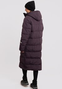 Lang, mørk lilla pufferjakke med hætte, der har vandret syninger og en sidelukning. Båret med sorte leggings og støvler.