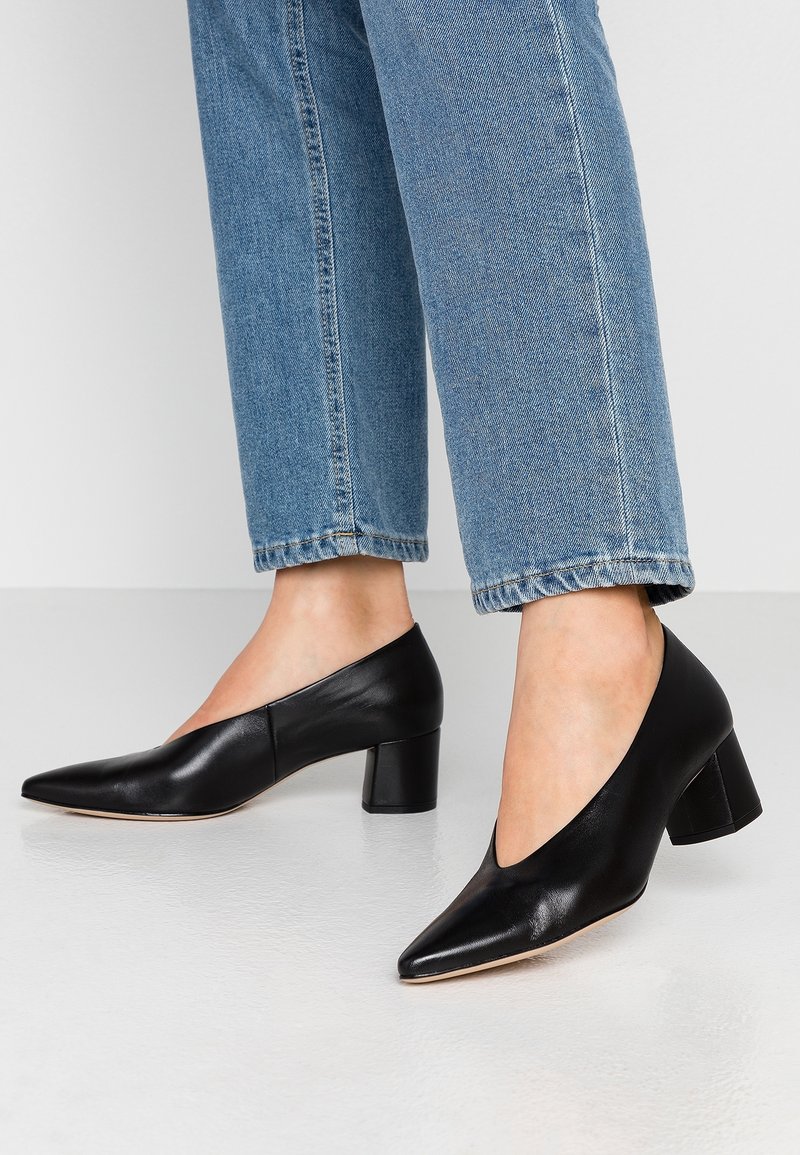 Personne portant des chaussures noires à talon bloc pointu associées à un jean bleu clair à jambes droites, debout sur un sol clair.