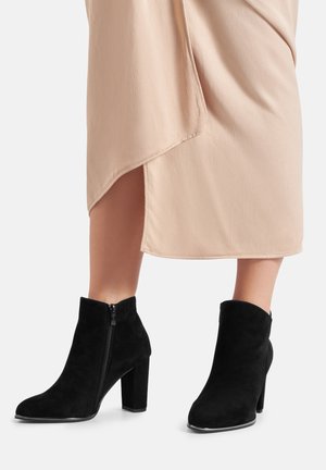 Czarne zamszowe botki za kostkę o gładkiej fakturze, z bocznym zamkiem i masywnymi wysokimi obcasami, zestawione z beżową spódnicą.