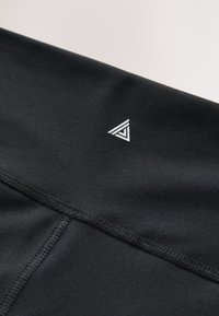 Tissu noir avec des coutures visibles et un petit logo triangulaire blanc composé de trois lignes imbriquées.