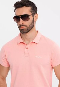 Ljusrosa polo med krage, med en två-knapps knäppning och en liten vit logotyp på bröstet. Modellen bär mörka solglasögon.